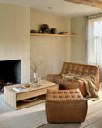 A cozy living room with tan leather furniture, an Ethnicraft Nordic Coffee Table in oak displaying books and pottery, a minimalist fireplace, neutral walls, decorative vases on a shelf, and sheer curtains letting in natural light.