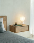 The Nordic II Hanging Bedside Table by Ethnicraft, crafted from solid European oak, is shown beside a bed with grey bedding. It's styled with a white sculptural lamp, two books, and a drink glass, set near sheer-curtained windows.