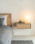 A modern bedroom with a solid European oak Ethnicraft Nordic bed frame, gray bedding, and an Ethnicraft Nordic II Hanging Bedside Table holding a glowing white lamp, two books, a candle, and eyeglasses against a light-colored wall.
