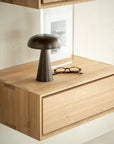 The Ethnicraft Nordic II Hanging Bedside Table in solid European oak displays a metallic mushroom lamp, tortoiseshell eyeglasses, and a framed black-and-white photo against a light-colored wall.