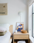 A modern, minimal bedroom features a solid European oak Ethnicraft Nordic bed and an Ethnicraft Nordic II Bedside Table with a candle, basket, and abstract art. Sunlight from the large window highlights a woven wall hanging on the white wall.