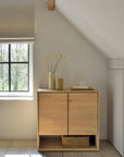 The Ethnicraft Nordic Sideboard - 2 door, crafted from solid oak, stands against a white wall beneath a sloped ceiling. Sunlight streams in as a beige vase with yellow flowers and a tray sit atop this piece from the Nordic Collection.