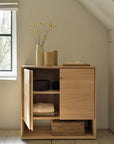 The Ethnicraft Nordic Sideboard - 2 door stands open to display folded blankets and baskets. On top sit a tall vase with yellow flowers, two books, a small stone, and a glass of water, all illuminated by sunlight from a nearby window.