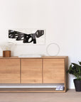 The Ethnicraft Nordic Sideboard - 4 door, made from European oak, stands against a white wall with abstract art above. On top sit a lamp, tray, and wire sculpture; beside and below are a potted plant and sneakers.