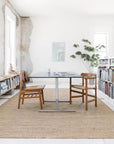 A bright, minimalist room featuring two Oakwood Collection chairs at a sleek table, bookshelves of books, a potted plant, and the Loloi Oakwood Rug in Wheat. Natural light illuminates textured walls through the windows.