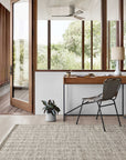 A modern home office with large windows, a wooden desk and chair, and a potted plant on the floor is enhanced by the Loloi Omen Rug - Grey, whose hand-woven chevron waves add style as sunlight streams in and green trees shine outside.