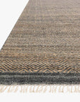 A close-up of the Loloi Omen Rug - Ink features hand-woven chevron waves in gray and brown, with fringe edging, shown at an angle on a white background.