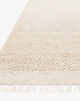The Omen Rug - Natural by Loloi is a beige, hand-woven jute rug with a subtle zigzag pattern and short fringes, displayed at an angle on a white background.