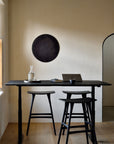 A modern minimalist workspace featuring an Ethnicraft black stained solid oak standing desk, two Ethnicraft Osso Barstools in Black, a glass carafe, a book, and a laptop. Round black wall art decorates the beige wall near an arched doorway filled with natural light.