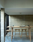 A minimalist dining area features a tall solid oak table and three Ethnicraft Osso Barstools in oak, accompanied by an open book and mugs, with large windows illuminating the textured walls and concrete floor.