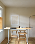 A minimalist workspace with European styling features a solid oak desk and two Ethnicraft Osso Barstools - Oak on a wooden floor, set against a cream wall with framed art and a small arched door, as sunlight streams through the window.