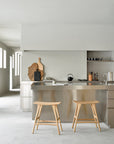 Minimalist modern kitchen with a stainless steel island, two Ethnicraft Osso Counter Stools in oak, light gray walls, and open shelves holding cups and utensils. Cutting boards and a teapot sit on the countertop as natural light fills the space.