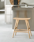 The Osso Counter Stool - Oak by Ethnicraft, featuring a curved seat, stands before a modern kitchen island with a stainless steel countertop. Minimalist decor and neutral tones complete the clean, contemporary look.