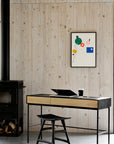 A modern workspace with an Ethnicraft wooden desk, an Osso Dining Stool in black by Ethnicraft, open laptop, and a glass of dark drink. Nearby are a black stained solid oak stove, abstract art, and stacked firewood against a wood-paneled wall.