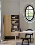 Minimalist home office with a light wood desk, black Osso Dining Stool by Ethnicraft, and shelving filled with books. A round window brings in natural light, neutral curtains hang to one side, and high ceilings with elegant moldings complete the look.