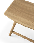 A close-up of the Ethnicraft Osso Dining Stool in oak shows its smooth, rectangular solid oak seat with rounded edges and part of its legs against a white background.