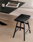 The Osso Dining Stool in black with a leather seat by Ethnicraft sits beside a dark table topped with an open magazine and small cup, set against light wood flooring and neutral decor for a refined European ambiance.