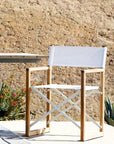 The Harbour Pacific Folding Chair - Teak, featuring a white seat and solid teak frame, is placed next to an outdoor table on a patio with a textured stone wall and green plants with red flowers in the background.