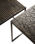 The Pentagon Nesting Coffee Tables - Whiskey by Ethnicraft feature metal frames and textured surfaces crafted from natural mineral powders: one table has a ridged, linear design while the other boasts a cracked, earth-like pattern with dark, metallic accents.