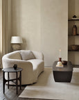 Minimalist living room featuring a curved cream sofa, side table, Ethnicraft PI Coffee Table in mahogany with candles and decor, neutral rug, large ceramic lamp, and shelves with pottery against light beige walls.