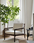 A cozy, modern armchair from the PI Collection with a mahogany frame sits by a window with white curtains. Nearby, an Ethnicraft PI Side Table - Mahogany holds a teapot and cup on the light wood floor next to a large leafy potted plant.