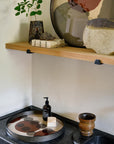 A black countertop with a wooden cup, soap dispenser, and Ethnicraft Pinot Layered Dots Glass Tray (Round/Small) sits below a wooden shelf holding a plant in a vase, stacked coasters, and ceramic decor against a light wall.