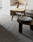 A minimalist room featuring a light wooden chair with woven seat, a dark wood coffee table, and the Armadillo&Co Pompeii Rug in Ore. A marbled ceramic bowl sits on the table against neutral-toned walls and floor.