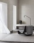 Minimalist room with light gray walls, sheer white curtains, a modern sculptural chair on the Armadillo&Co Pompeii Rug in Rye, a narrow bench along the wall, plus a black floor lamp and vase.
