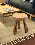 The Priam Side Table by Noir Trading Inc., a small solid teak table (15 x 15 x 18.5) with three angled legs, sits on a textured cream rug near a light wood coffee table, in front of a dark gray sofa.