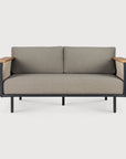 The Corbey Outdoor Sofa by Ethnicraft is a stylish two-seater with grey cushions, a black metal frame, and light wood armrests—perfect for your outdoor space against a white backdrop.