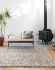 A bright, minimalist living room features a light beige sofa, patterned cushion, potted plant, wood-burning stove, and the hand-knotted 100% wool Quarry Rug - Stone by Loloi on wooden floors as sunlight streams in.