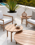 A modern patio featuring Ethnicrafts Quatro Outdoor Coffee Table in teak, surrounded by cushioned wooden chairs, a potted succulent, a decorative lantern, and a snack bowl—an inviting setting amid lush greenery.