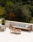 A modern outdoor patio features Ethnicraft light wood furniture, gray cushions, the Quatro Outdoor Coffee Table in teak by Ethnicraft, a potted plant, and trees in the background on a sunny day.