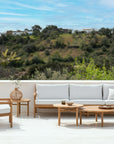 Outdoor patio with Ethnicraft wooden furniture, including a teak sofa, armchair, and the Quatro Outdoor Coffee Table - Teak by Ethnicraft. The patio overlooks a scenic green hillside under a blue sky with scattered clouds.