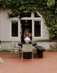 A stylish patio features a black table with Moes Silla Outdoor Dining Chairs (set of 2) and a bench on red tiles beside a vine-covered building; the table is topped with a white vase of flowers, and a white metal staircase stands to the left.