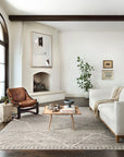Bright, modern living room with neutral tones—a cream sofa, brown leather chair, wooden coffee table, fireplace, large abstract art, indoor plant, and the Loloi Rayan Rug in Ash on dark wood floors. Light streams through a large arched window.