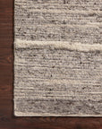 A close-up of the Loloi Rayan Rug - Charcoal/Ivory shows its thick, chunky yarns and subtle striped pattern near the edge, displayed on a dark wooden floor in soft neutral shades.