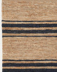 The River Rug - Natural & Indigo by Armadillo&Co is a Fair Trade hand-woven tan jute rug with horizontal indigo stripes of varying thicknesses. Only a partial view is displayed on a white background.