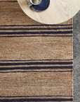 The Armadillo&Co River Rug - Natural & Indigo, featuring hand-woven horizontal navy stripes, is laid on a textured floor. In the upper corner, a round light wood table displays a gray cup with a dark blue saucer.