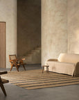 A minimalist living room with textured beige walls, a beige sofa, a wooden chair, and a small stool features the Armadillo&Co River Rug in Natural & Indigo on the concrete floor. Stairs are visible in the background.