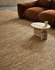 A cozy living room features the Armadillo&Co River Rug - Natural, a low marble coffee table with a cup atop it, and a plush brown sofa set against a light-colored wall.