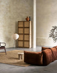 A minimalist living room features a brown sectional sofa, wooden armchair, woven cabinet, round floor lamp, small round table, and an Armadillo&Co River Rug - Natural adding texture beneath a decorative branch in a vase against beige textured walls.