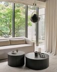 A modern living room with large windows, a beige sofa, cream curtains, and two round black Roller Max Coffee Tables by Ethnicraft. Sunlight streams in from outside greenery while a sleek black pendant light hangs above.