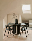 Minimalist dining area featuring the Ethnicraft Roller Max Round Dining Table in dark mahogany, four black chairs, a gray vase with dried branches, light wooden floor, beige walls, skylight, cream cabinet, and a light sofa in the background.
