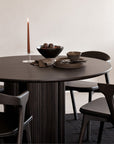The Ethnicraft Roller Max Round Dining Table in dark mahogany is styled with matching chairs, stacked plates, a bowl, cup, folded napkins, fruit bowl centerpiece, and a tall lit candle on a glass holder.