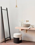 Minimalist bathroom with a black-framed floor mirror, floating light wood vanity, round vessel sink, gold faucet, small vase of flowers, pendant light, and the Ethnicraft Roller Max Pouf featuring a textured black mahogany base.