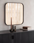 A lit brown candle in a geometric holder sits on the Ethnicraft Roller Max Sideboard - 88, next to a closed book, beneath a large square mirror that reflects vertical wood patterns.