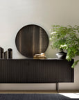 The Ethnicraft Roller Max Sideboard - 88, with a sleek black teak finish and adjustable shelves, stands against a light wall, styled with a large round mirror, minimalist black vases, stacked books, and a leafy potted plant in a bright interior.