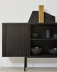 The Ethnicraft Roller Max Sideboard - 88, featuring black wood with vertical grooves, stands against a white wall. Stacked books and a dark object sit on top, while adjustable shelves inside hold black ceramic bowls and a matching pitcher.