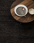 A round wooden table with a ceramic plate, small bowl, and bronze spoon sits atop the Armadillo&Co Savannah Rug - Carbon, blending inviting texture and expert craftsmanship.
